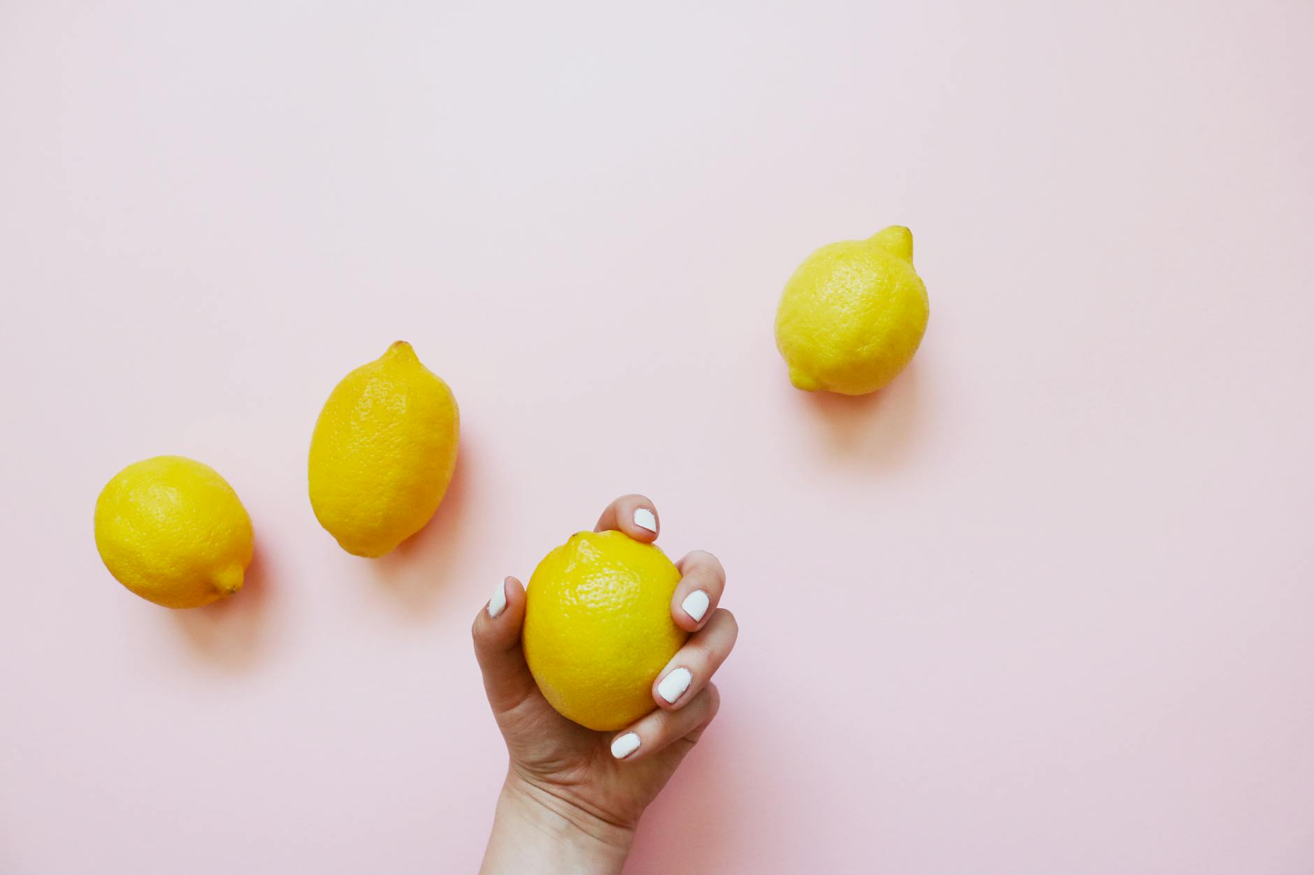 A hand with white nails holding a ripe lemon on soft pink background, surrounded by fresh lemons