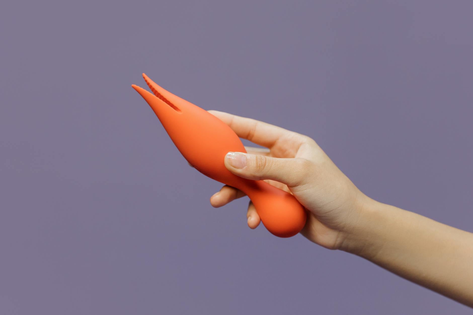 A hand holding a sleek orange lemon vibrator against a minimalist purple backdrop, representing modern intimacy and openness