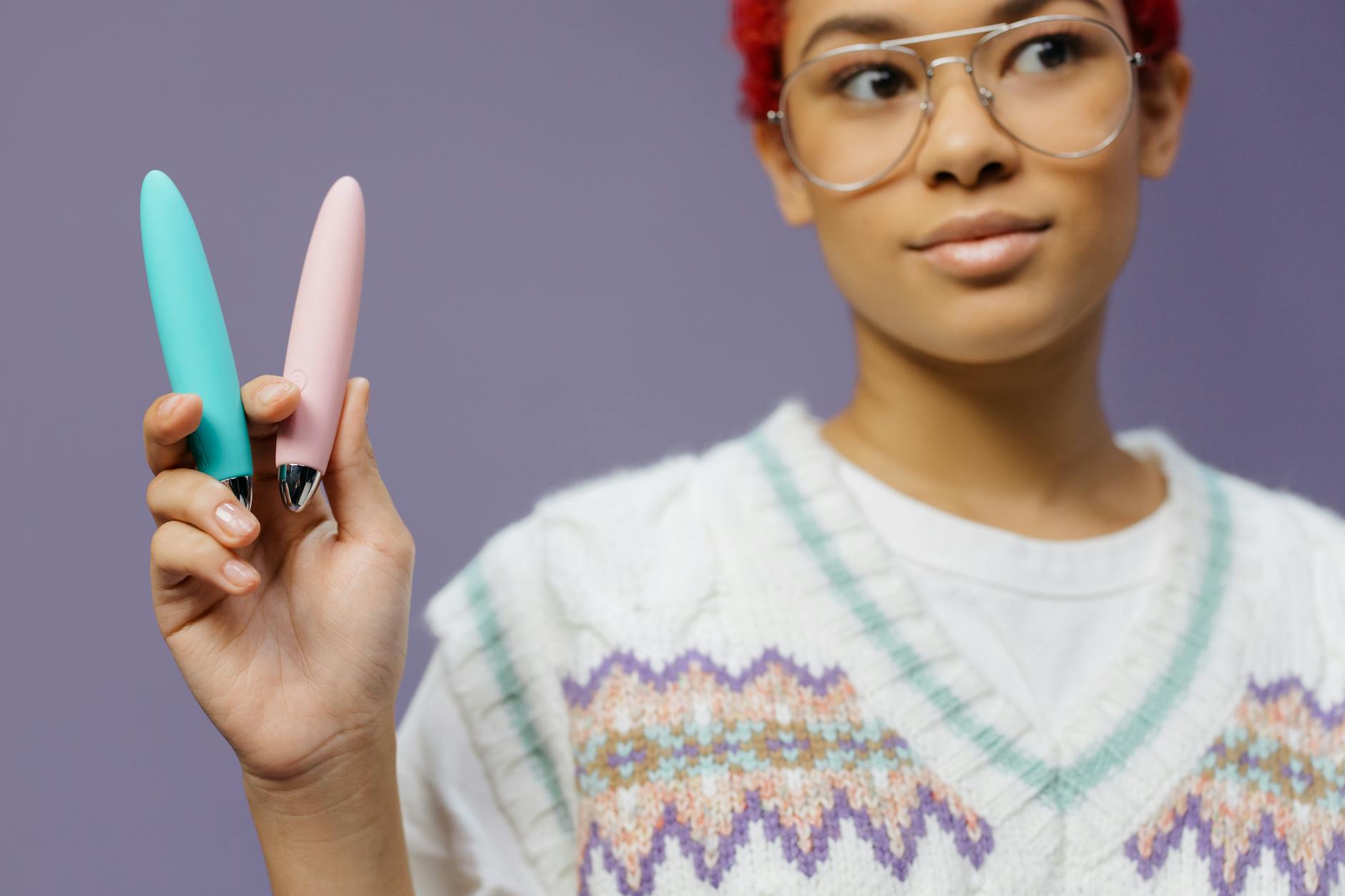 A woman with glasses holding colorful vibrators against a purple background.