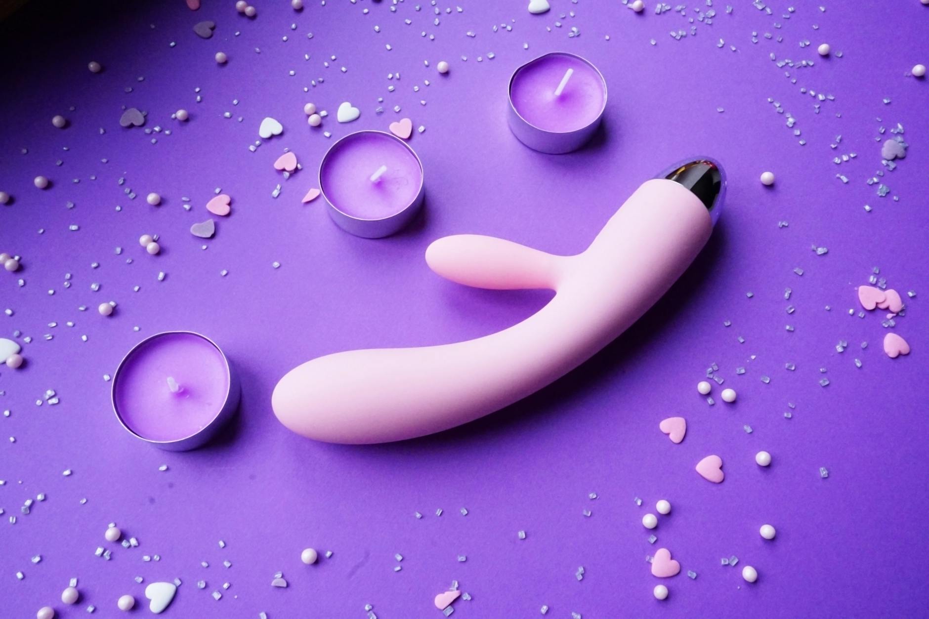 Pink vibrator surrounded by candles and heart confetti on a purple surface, representing intimate pleasure and recovery